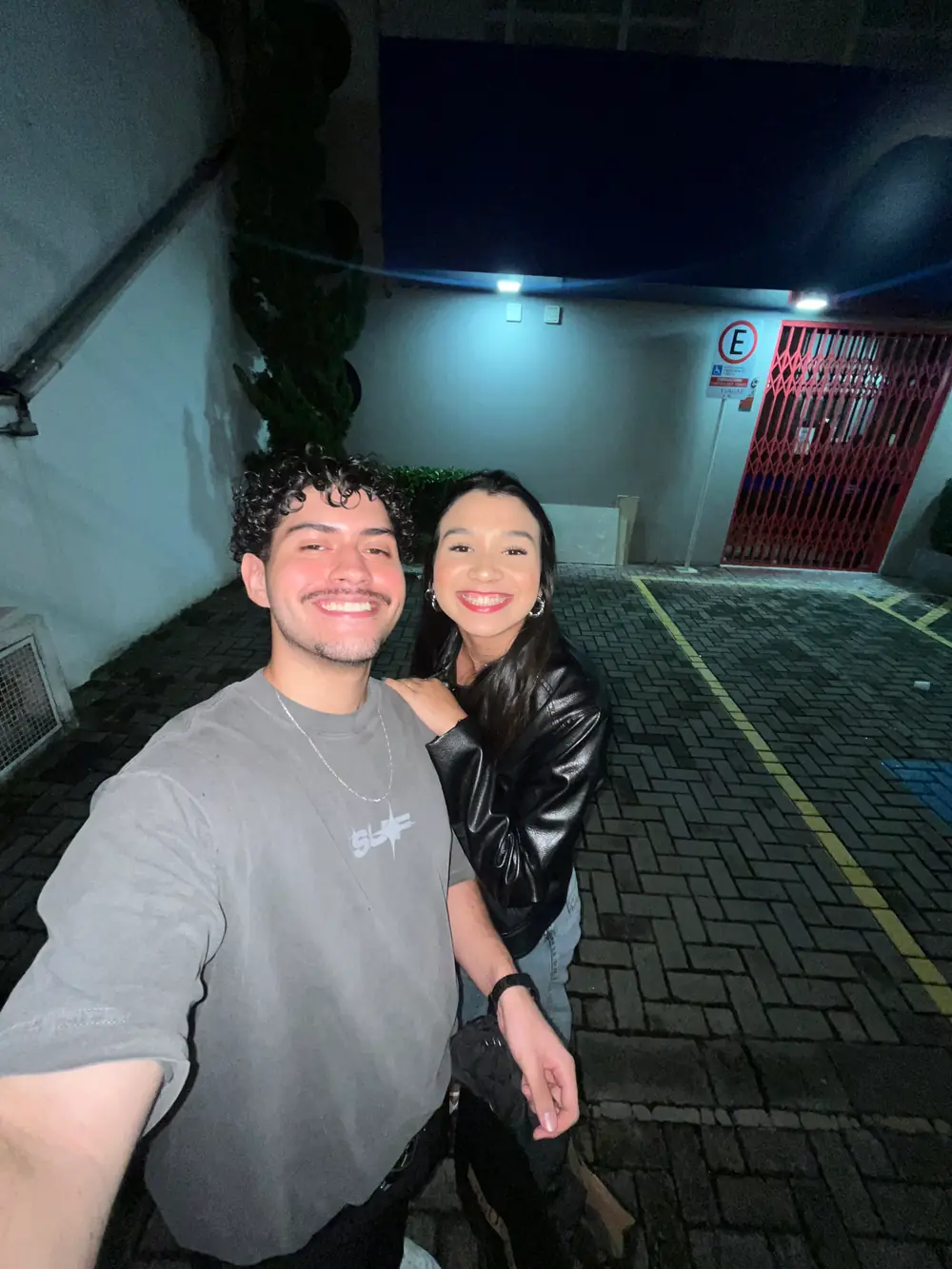 Selfie na calçada à noite, ela de jaqueta de couro